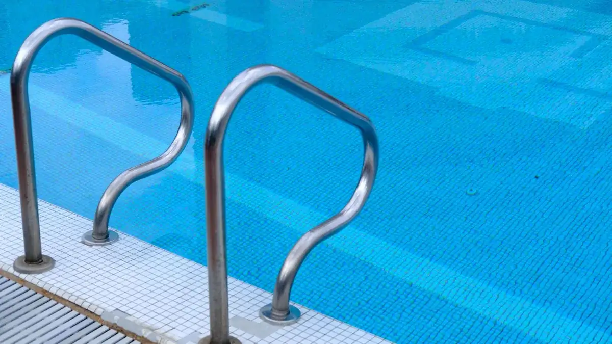 Barras de acero inoxidable de una escalerilla de piscina ancladas al borde alicatado, entrando en el agua azul.