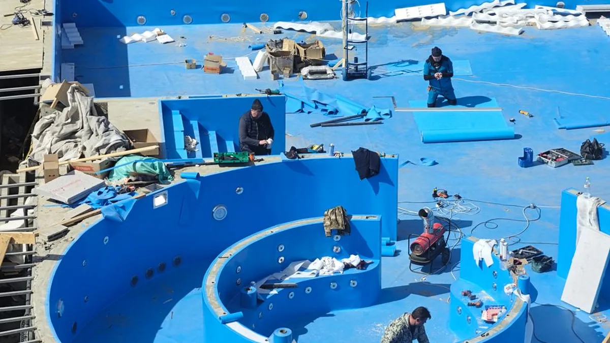 Piscina en obra vista desde arriba, con el vaso ya impermeabilizado en azul, una zona circular y varios operarios trabajando entre herramientas y materiales.