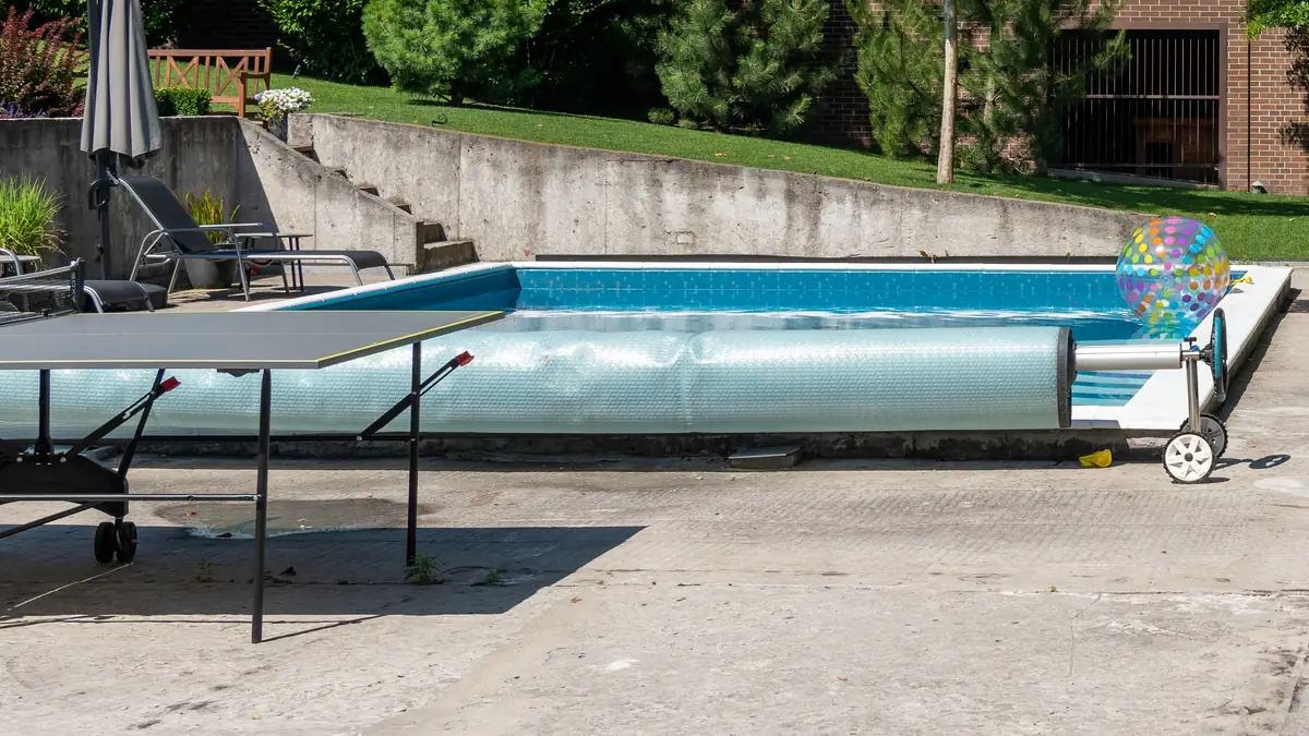 Piscina en un jardín con el cobertor de burbujas enrollado en un enrollador con ruedas junto al borde, y una mesa de ping-pong en primer plano.