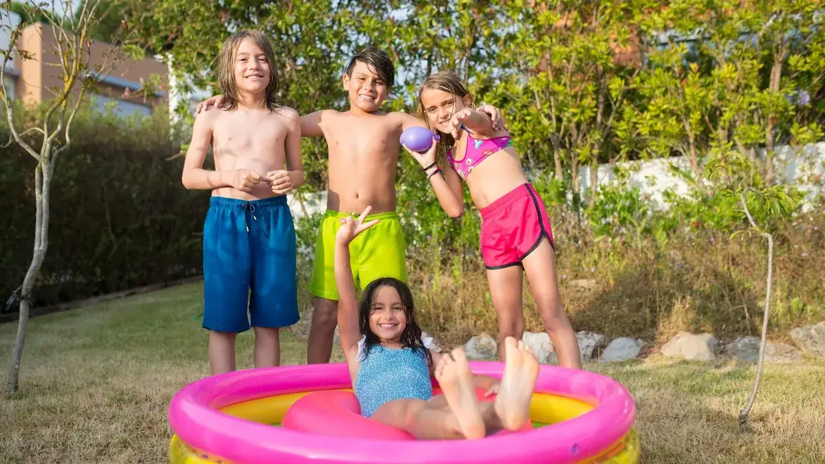Cuatro niños en un jardín junto a una piscina hinchable redonda de colores; una niña está sentada dentro.