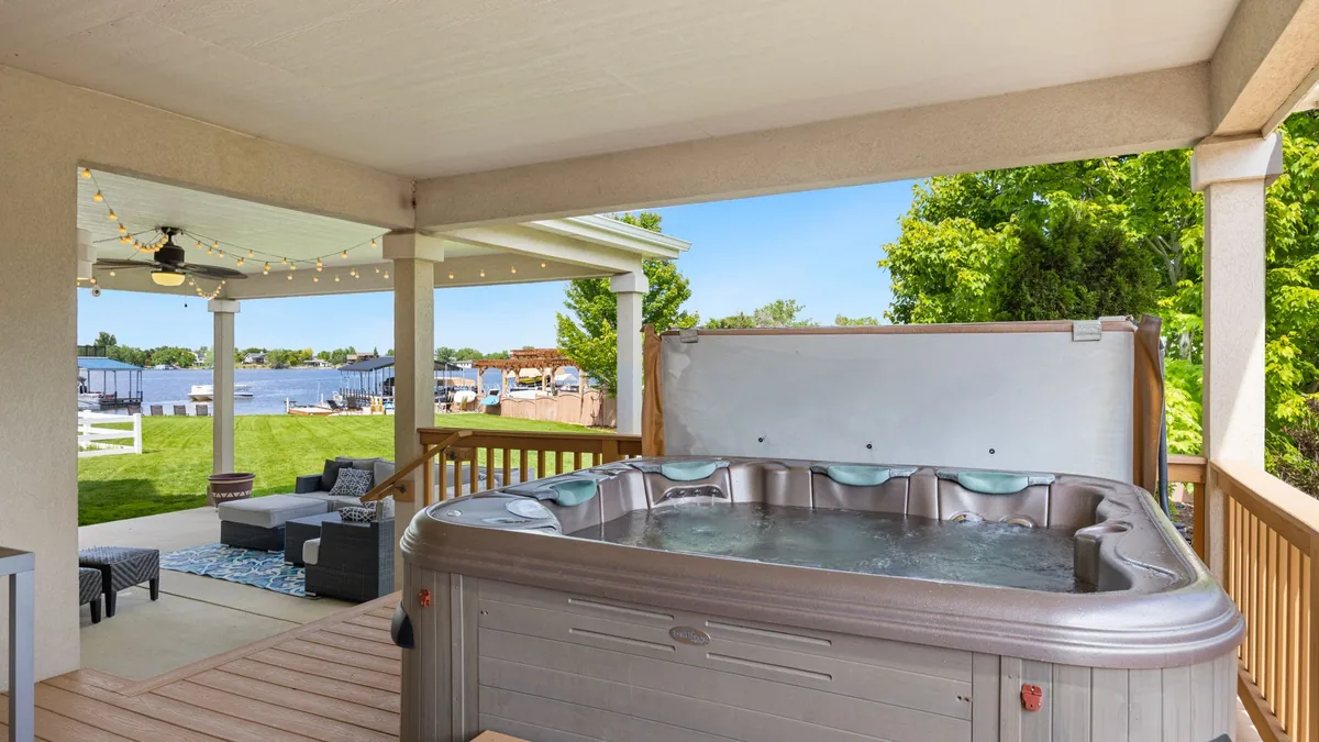 Jacuzzi portátil en una terraza cubierta, con la tapa apoyada detrás y vistas a un lago con embarcaciones.
